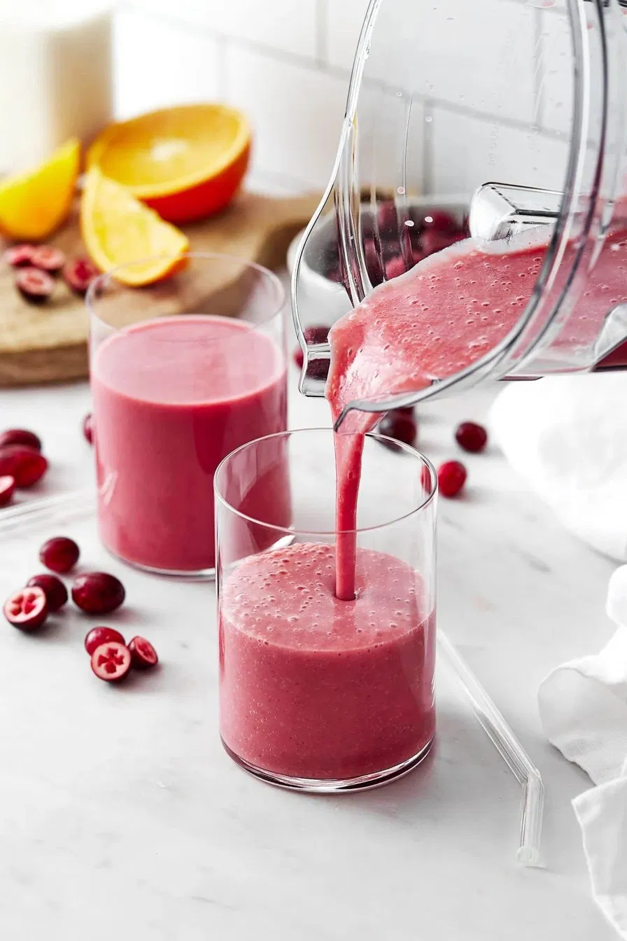 Pink smoothie being poured from a blender into a glass with cranberries and orange slices